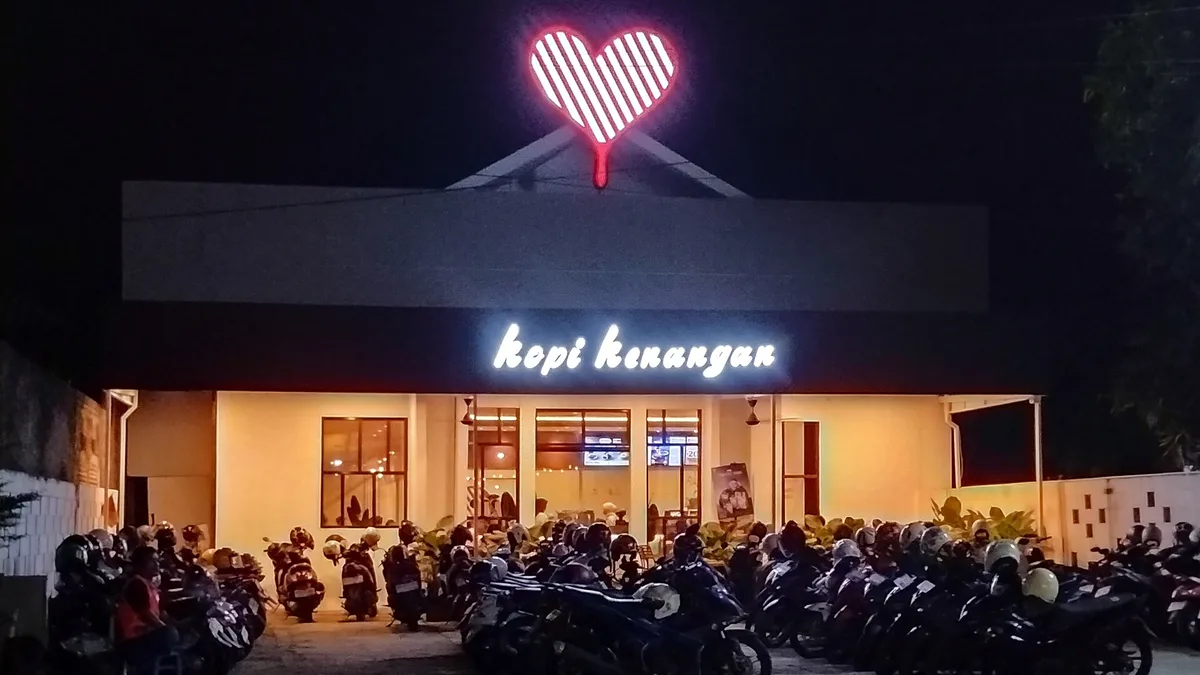 Gerai Kopi Kenangan di Ngawi pada malam hari: simbol hati bergaris menyala merah di atap dan wordmark kopi kenangan berlampu putih di atas pintu masuk
