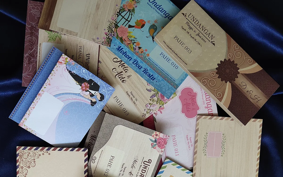 Foto belasan undangan pernikahan blangko cetak dari Indonesia bermotif kayu, floral, burung, dan amplop bergaris ala surat pos, ditata di atas kain satin biru