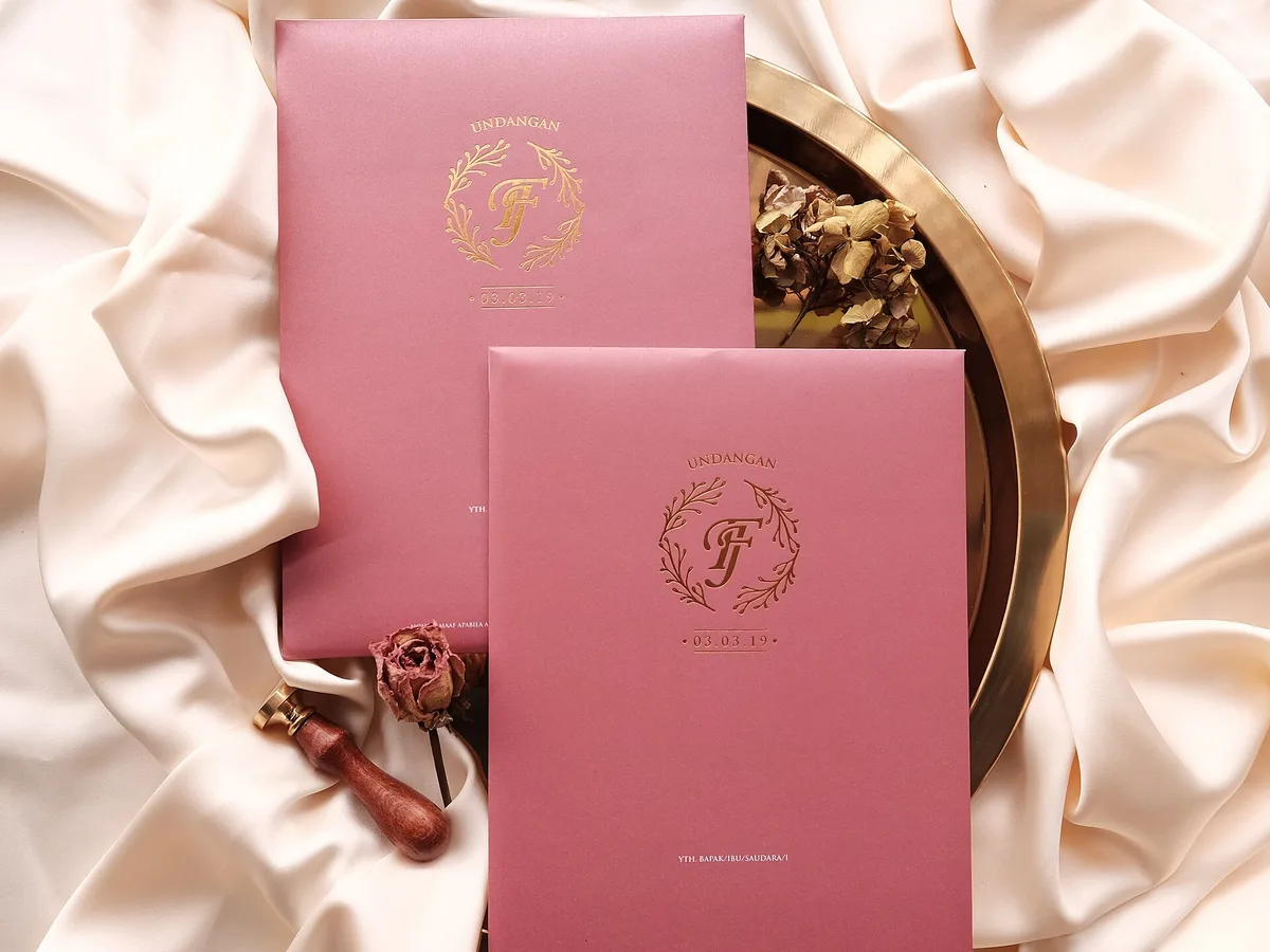 Dua undangan pernikahan custom berwarna dusty pink dengan monogram dan bingkai daun gold foil, di atas nampan kuningan dan kain satin bersama stempel lilin